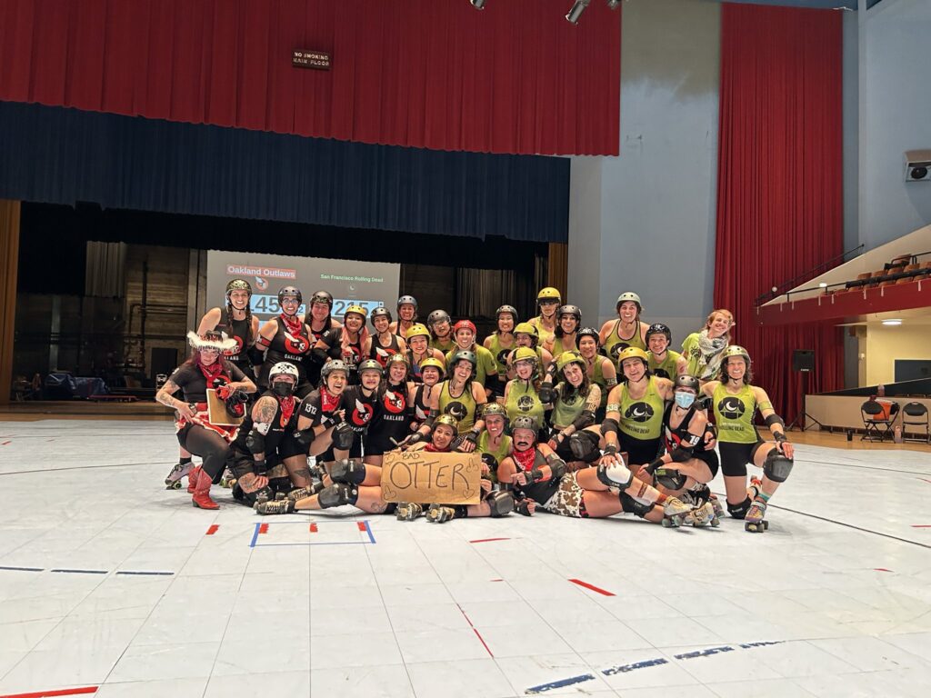 Skaters from the Oakland Outlaws and SF Rolling Dead pose for a team photo in front of the scoreboard holding a sign reading "Otter"
