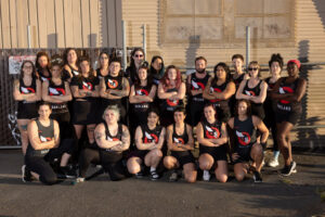 Team photo of people in Oakland Outlaws jerseys (black shirts with a skull wearing a winged helmet) in front of a corrugated metal building.