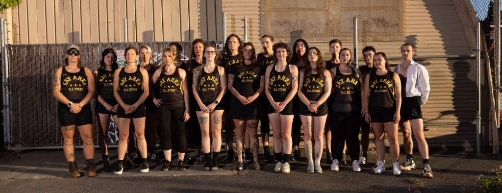 A group of skaters wearing black jerseys with "Bay Area All Stars" printed on them in gold and black shorts stands in front of a metal, industrial building.