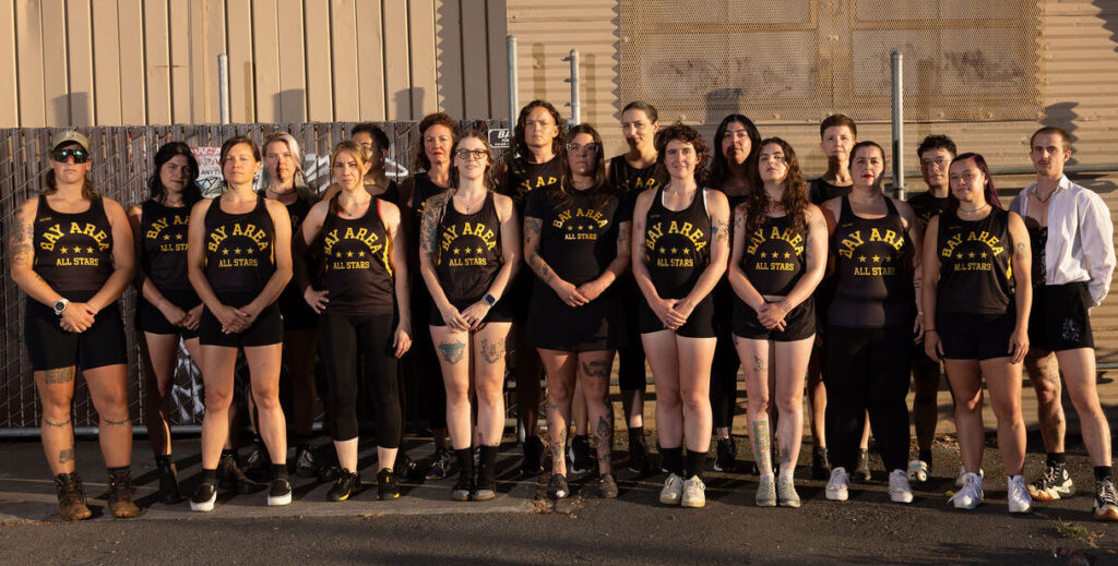 A group of skaters wearing black jerseys with "Bay Area All Stars" printed on them in gold and black shorts stands in front of a metal, industrial building.